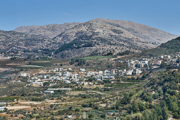 A panoramic view of a rural town nestled between rugged mountains and green agricultural fields on a sunny day.