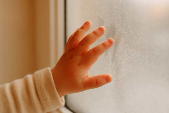 Curious child's hand touching frosted glass window indoors, exploring texture and temperature in natural soft daylight