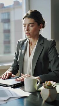 A businesswoman in a suit sits at her desk, looking stressed as she stares at her laptop, then rubs her temples and looks up thoughtfully, vertical footage