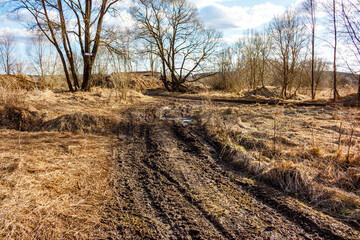 A gnarly, muddy dirt track cuts through withered grass and bare trees under a bright sky, hinting at tough off-road journey