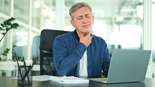 Distressed businessman, dressed in a blue denim shirt and white t-shirt, experiences painful sore throat in a modern office. Uncomfortable male stops typing on laptop, feeling unwell while at work.