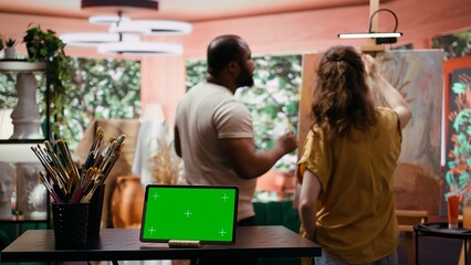 Two artists painting on a canvas next to green screen on tablet display, working together on their new masterpiece for fine art gallery exhibition. Diverse team of painters in art studio. Camera B.
