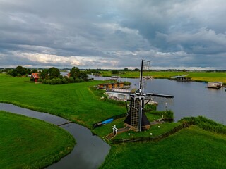 Aerial view of Dutch countryside black and white windmill by winding canal, green fields, boats, stilted houses, and dramatic cloudy sky create a serene and picturesque rural scene.
