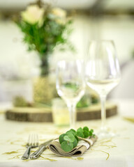 Eine festlich gedeckte Tafel in einem hellen Festzelt. Das Bild zeigt einen runden Tisch, der mit einem weißen Tischtuch und einem stilvollen gold-grünen Tischläufer geschmückt ist. Im Zentrum steht e