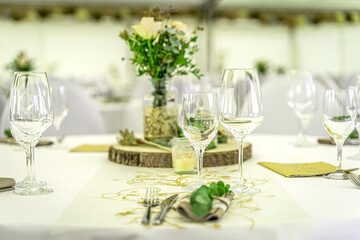 Eine festlich gedeckte Tafel in einem hellen Festzelt. Das Bild zeigt einen runden Tisch, der mit einem weißen Tischtuch und einem stilvollen gold-grünen Tischläufer geschmückt ist. Im Zentrum steht e