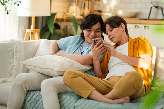 Asian male couple smiling and relaxing together on a cozy sofa, browsing social media or shopping online on a smartphone.Concept of LGBTQ+ relationship, domestic life and digital connection. - Powered by Adobe