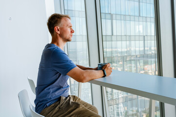A young man is sitting at a table and drinking coffee looking out the window at skyscrapers and business centers