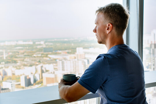 A young man is sitting at a table and drinking coffee looking out the window at skyscrapers and business centers - Powered by Adobe