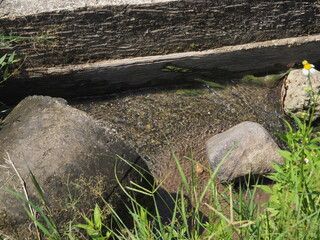 A close-up view of clear, shallow water flowing over pebbles in a narrow channel between two large, rounded concrete blocks and beneath a rough concrete slab, surrounded by green grass.