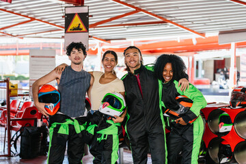 Obraz premium Group of smiling diverse friends in racing suits and holding helmets, ready to race go karts on a track