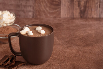 Cozy hot chocolate or sweet homemade cocoa drink served in mug or cup decorated with sugar marshmallows on dark brown table with copy space with chocolate and powder ingredient for winter breakfast