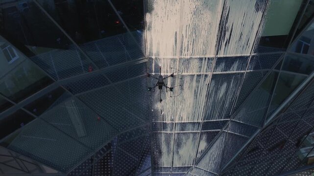 aerial view of Industrial drone spraying foam and water while cleaning glass facade of modern office building with sunset reflections