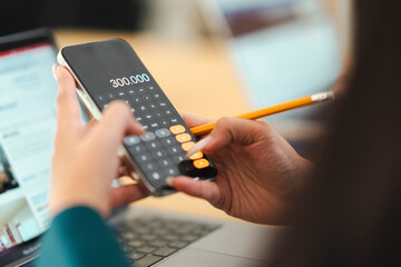 A person uses a smartphone calculator to input numbers, with a laptop and pencil nearby, suggesting financial planning or data analysis.