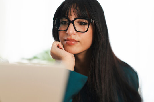 A young woman with glasses rests her chin on her hand while looking at a laptop screen, deep in thought.