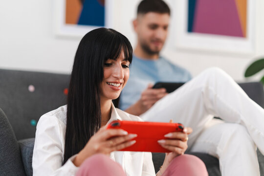 A young woman smiles as she plays a handheld video game console, while a man in the background also uses a similar device.