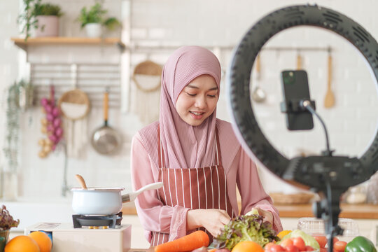 young beautiful muslim woman in hijab prepare organic fruits and vegetables for cooking on video live streaming in the kitchen,concept healthy food,technology,new marketing