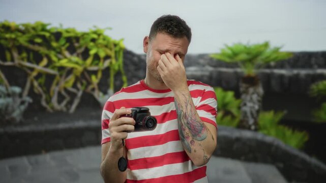 Young man holding camera outdoors with plants in the background, expressing concern, wearing striped shirt, tattooed arm visible, suggesting emotional moment.