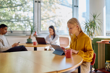 Blonde woman using a digital tablet at a coworking desk, with diverse colleagues working in the sunny background