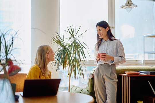 Two women colleagues smiling, listening, and talking during a casual coffee break in a bright, modern office space