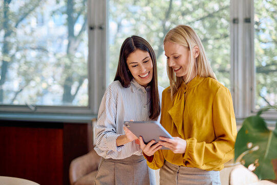 Two smiling businesswomen discussing work and sharing ideas while looking at a digital tablet at the modern office