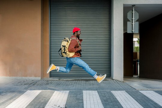 Cheerful man in casual clothes jumping with backpack on urban street