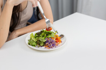 Close-up of Asian woman eating fresh vegetable salad with fork at a white table. Concept for healthy eating, weight control, clean food, vegan lifestyle, and nutrition awareness.