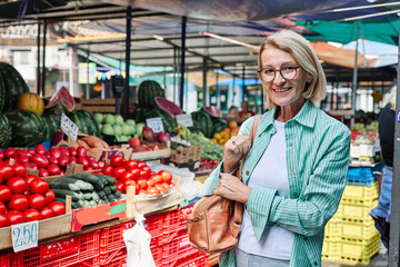 A woman enjoys selecting ripe tomatoes at a bustling outdoor farmers market. Colorful fruits and vegetables surround her as she smiles at the freshness of the produce