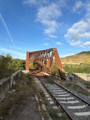 An old, rusty iron railway bridge spans a valley surrounded by nature, amidst golden hills and a clear sky.