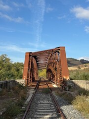 An old, rusty iron railway bridge spans a valley surrounded by nature, amidst golden hills and a clear sky.