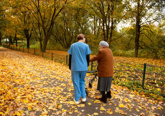 Obraz premium Elderly woman with walker taking a walk through the park with fallen leaves with help of caretaker or nurse in blue uniform