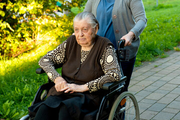 Caretaker in blue uniform taking a walk with elderly female patient through park with help of...
