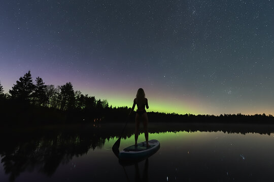 Adventurous woman on a stand-up paddleboard floats on a tranquil Estonian lake, gazing at the magical Aurora Borealis and stars. - Powered by Adobe