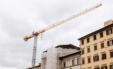 Yellow tower crane extending over residential buildings under renovation with scaffolding and safety netting against overcast sky in urban Italian city