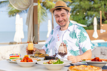 Man enjoying tropical outdoor dining by pool, wearing summer shirt and hat, surrounded by colorful drinks and food, tropical ambiance, vacation relaxation concept.