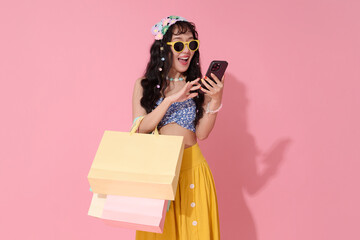 Trendy young asian woman holding shopping bags using smartphone isolated on pink background expressing excitement for online shopping deals and digital lifestyle happiness