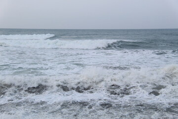 Ocean waves crashing on the shore under a cloudy sky. Blue sea water with white foam shows the power and beauty of nature, perfect for themes of travel, nature, energy, and environment