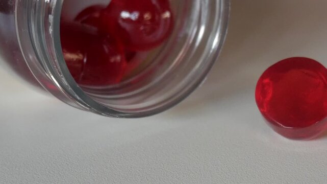 Close-up of red vitamin gummies spilling from a glass jar on a white surface under soft light.