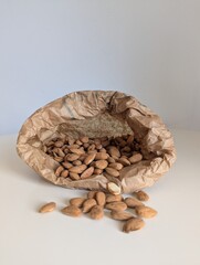Raw almonds pouring from a brown craft paper bag onto a bright white surface. Capturing natural, healthy movement.
