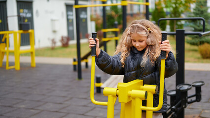 Obraz premium girl playing on outdoor exercise equipment in park setting. wearing puffy jacket on chilly day, exploring fitness playground area. engaging activity for active childhood development