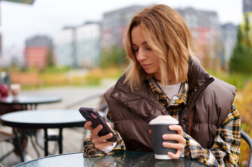 woman using smartphone at outdoor café, holding coffee cup, enjoying casual autumn atmosphere....