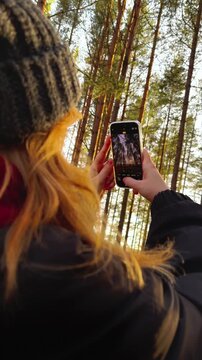 Close-up shot from behind of a woman wearing a dark beanie and jacket holding a smartphone to capture a vertical video of the tall pine trees and golden sunlight streaming through the autumn forest 
