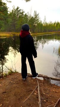 Full shot from a low angle shows a woman wearing a dark jacket and beanie standing on a small, exposed dirt hill overlooking a calm forest lake during a cloudy autumn evening, with exposed tree roots 
