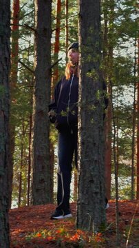 Medium shot of a woman with a dark jacket and beanie walking on a mossy hill through a pine forest during a golden hour sunset, with warm natural light filtering through the tall trees