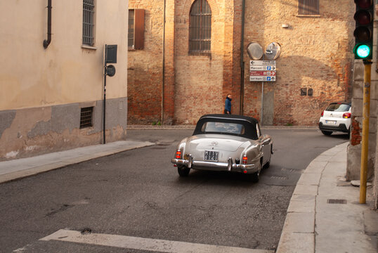 Vintage Mercedes-Benz 190 SL convertible cruising a sunny narrow European street past brick buildings and traffic lights, open roof freedom