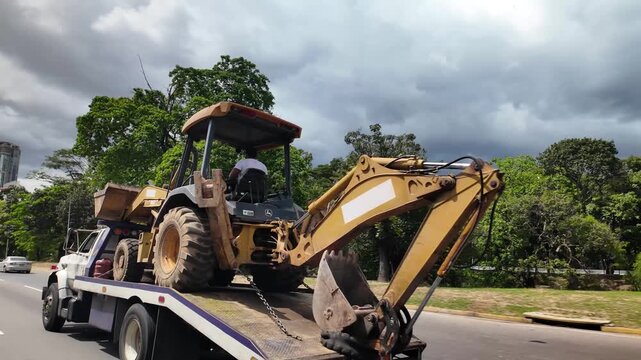 yellow backhoe loader being transported on a flatbed truck driving along a highway surrounded by green trees and cloudy sky