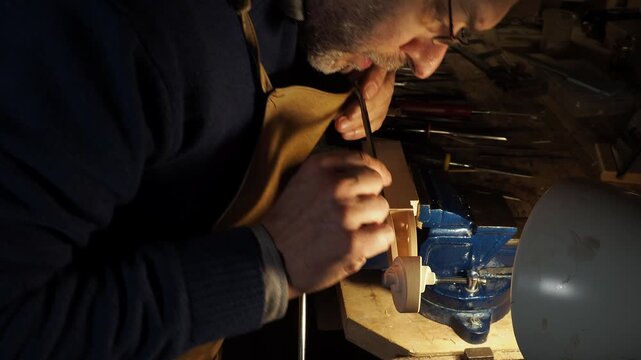 Luthier leans over workbench, carefully carving violin scroll clamped securely in a vise, With steady hands and a sharp tool, he refines the curves and details, demonstrating traditional craftsmanship