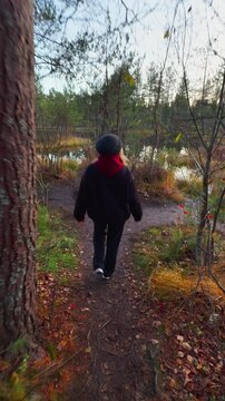 Full shot from a low angle shows a woman wearing a dark jacket and beanie standing on a small, exposed dirt hill overlooking a calm forest lake during a cloudy autumn evening, with exposed tree roots 