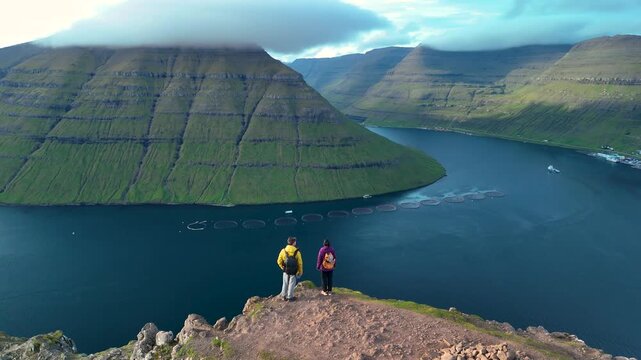 Breathtaking view from the summit shows two hikers gazing at the lush cliffs and calm bay in Klakkur Faroe Islands. The landscape showcases natures beauty during a clear day.