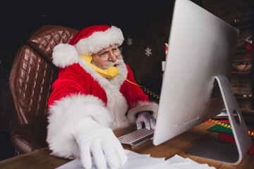 Santa at desk working on computer with yellow phone in a cozy home workshop full of holiday...