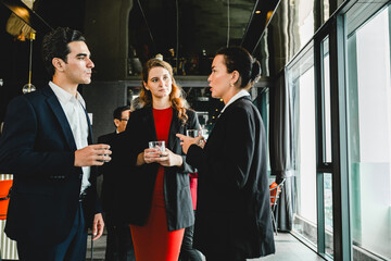 A group of professionals in suits networking and chatting with drinks in a modern lounge with large windows, capturing a corporate social event and casual business conversation.
